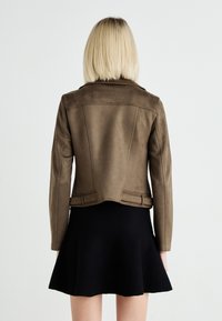 Bruine faux leren jas met een getailleerd ontwerp en naden aan de achterkant, gedragen over een zwarte flared rok.