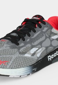 Γκρι και μαύρο παπούτσι τρεξίματος Reebok με κόκκινες λεπτομέρειες, μαύρα κορδόνια, σχέδιο από πλεκτό διχτυωτό υλικό και απορροφητικό μαξιλάρι Superfloat στη σόλα.