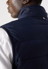 Temno modri napihnjen jopič z visokim ovratnikom, prešito zasnovo in subtilnim horizontalnim šivanjem. Vključuje kontrastne navpične stripe.