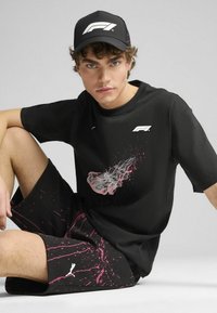 Sort t-shirt med hvidt logo og pink grafik, parret med sorte shorts med pink sprøjtedesign. Sort kasket med logo akcenter.