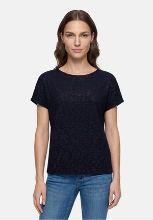 Kvinde med brunt hår iført en kortærmet sort tekstureret top og blå jeans, stående mod en enkel lys baggrund.
