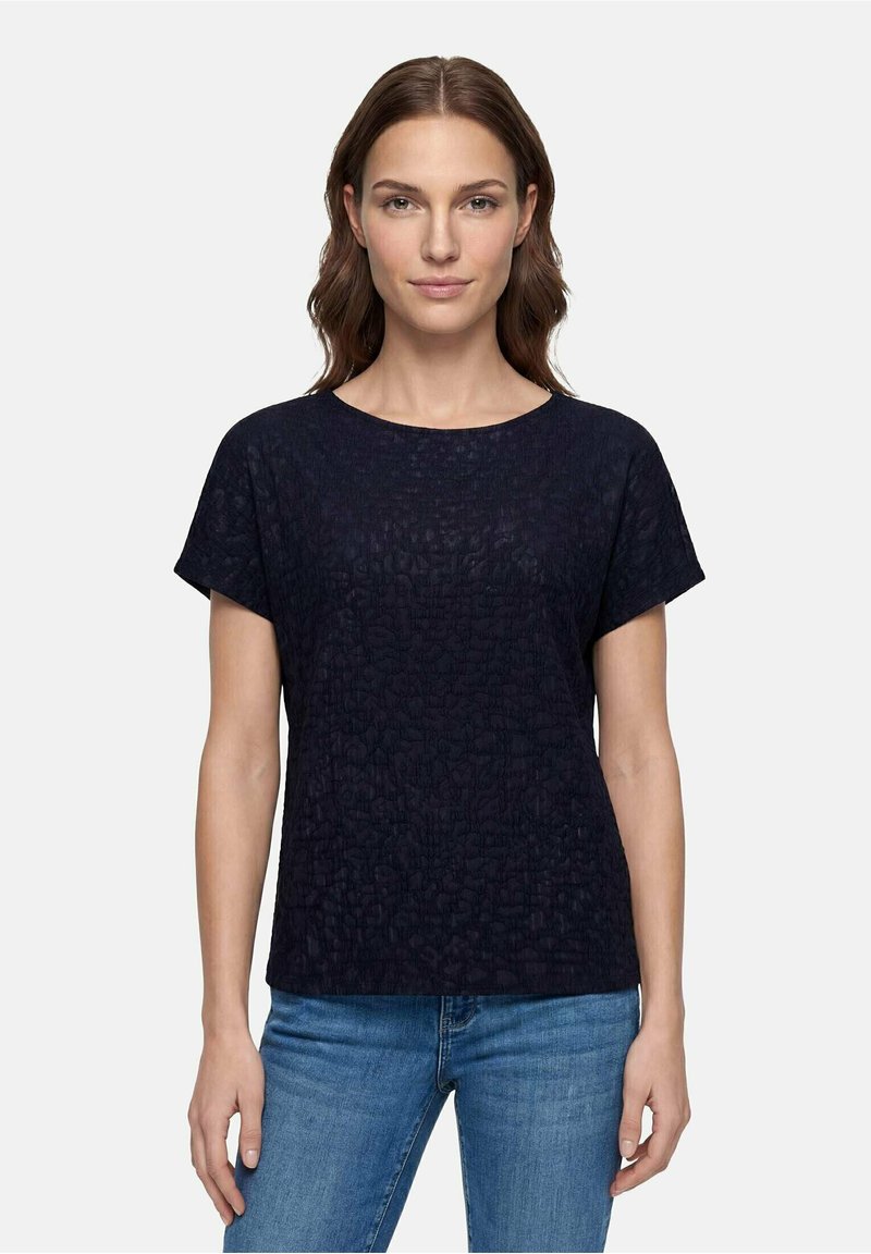Femme aux cheveux bruns portant un haut noir texturé à manches courtes et un jean bleu, debout devant un fond clair uni.