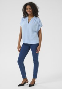 Blouse bleu clair à manches courtes avec un décolleté en V et une coupe loose, associée à un jeans foncé et des escarpins noirs à bout pointu. Texture du tissu lisse.