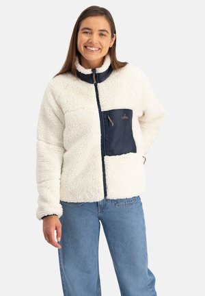 Glunderende vrouw die een wit fleecejack met een marineblauwe voorkantzak en lichtblauwe spijkerbroek draagt, staande tegen een witte achtergrond.