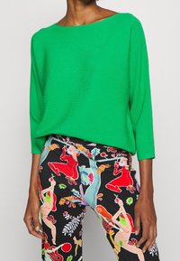 Zelený žebrovaný svetr s širokým výstřihem a dlouhými rukávy, doplněný černými kalhotami s barevným uměleckým vzorem postav a rostlin.