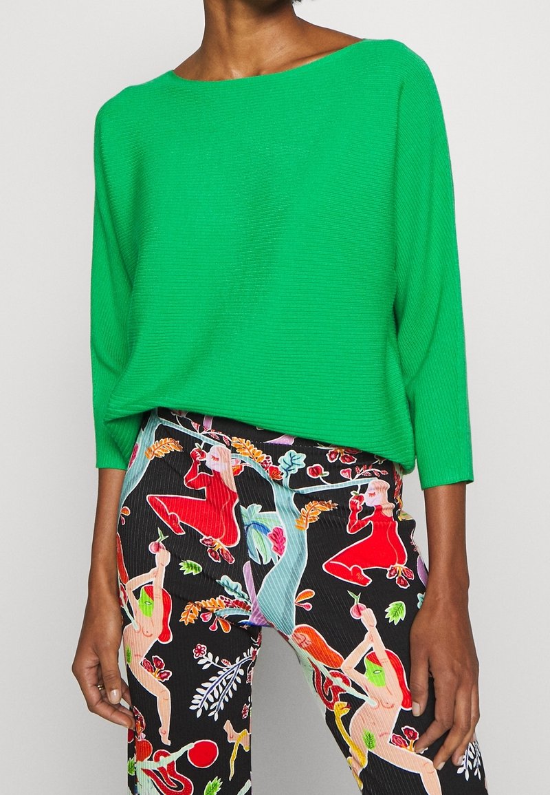 Zelený žebrovaný svetr s širokým výstřihem a dlouhými rukávy, doplněný černými kalhotami s barevným uměleckým vzorem postav a rostlin.