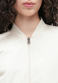 Blouson aviateur beige clair avec fermeture éclair à l'avant, col côtelé et texture lisse. Présente un design minimaliste avec des coutures discrètes.