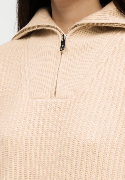 Gros plan sur un pull beige en tricot avec une fermeture éclair métallique à moitié descellée et une texture côtelée sur la partie supérieure de la poitrine et du cou d'une personne.