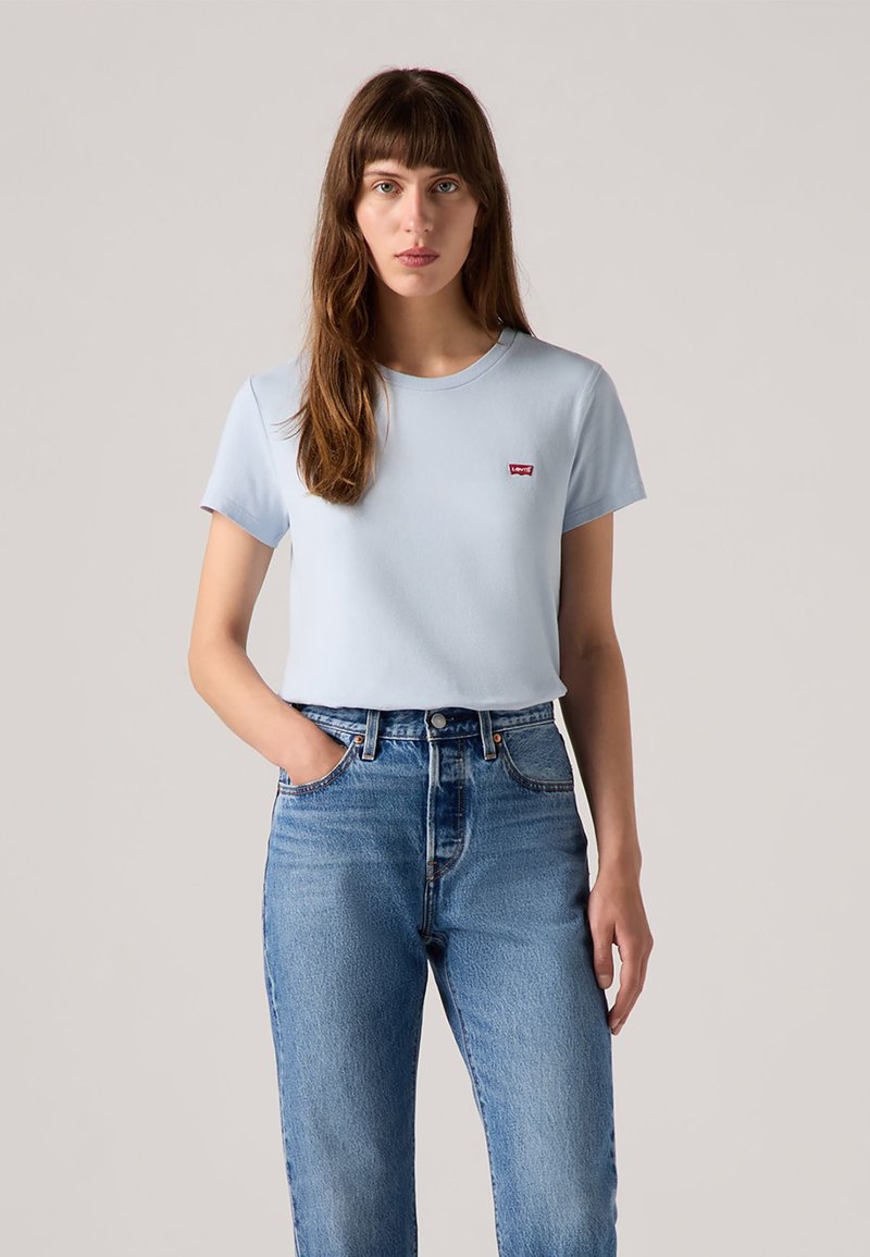 Camiseta de manga corta azul claro con escote redondo y un pequeño detalle de logo rojo en el pecho, combinada con unos jeans de talle alto azules.