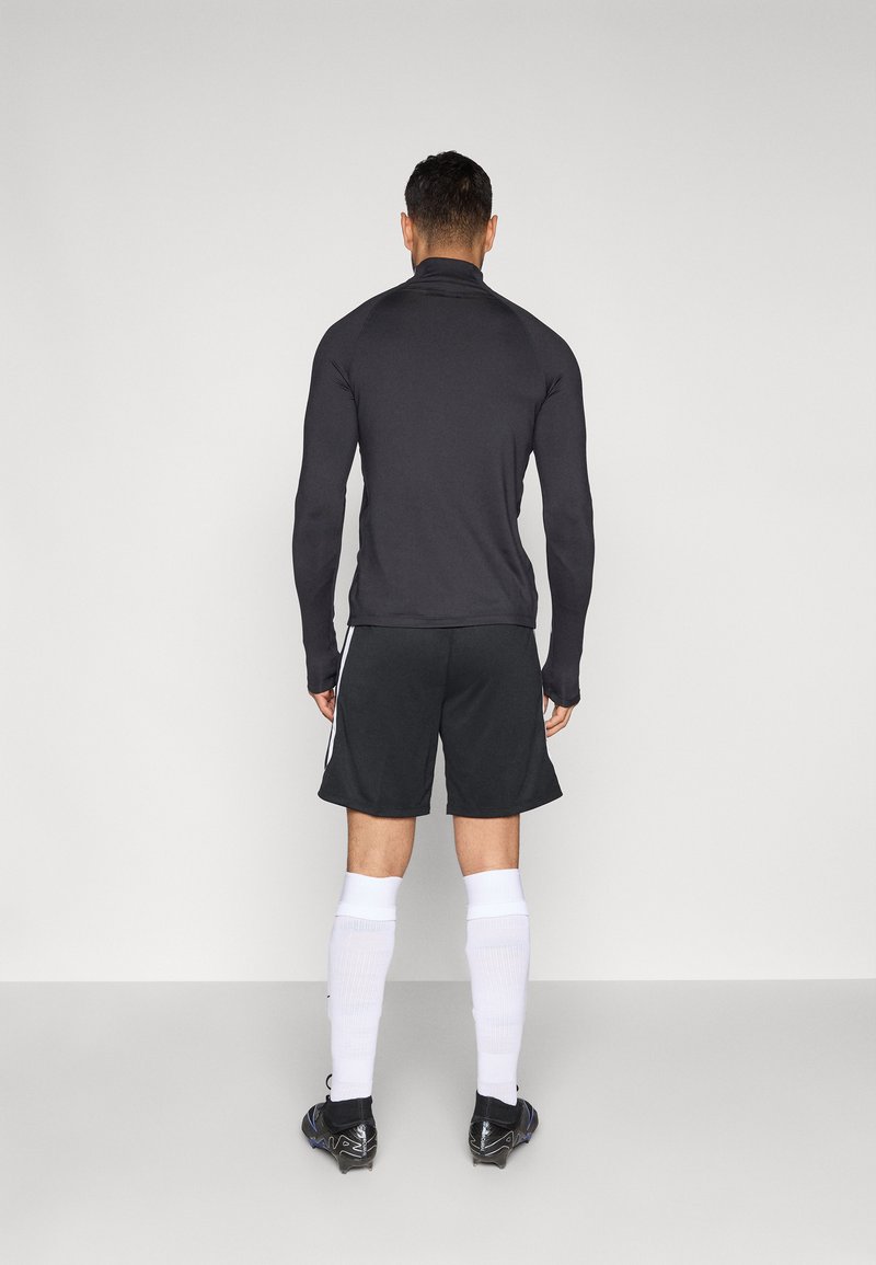 Sportliches Outfit mit einem enganliegenden schwarzen Langarmshirt und schwarzen Shorts mit weißen Seitenstreifen, kombiniert mit weißen Socken und schwarzen Stollenschuhen.