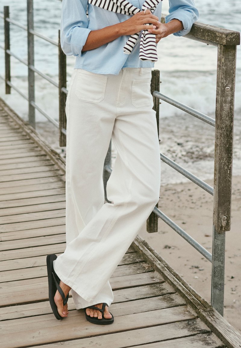 Personne portant un pantalon blanc ample, une chemise bleu clair et des sandales noires, se tient sur une jetée en bois au bord de la mer, tenant un pull rayé.