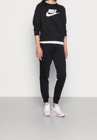 Svart Nike-tröja med vit logotyp, tillsammans med svarta joggingbyxor. Skorna är vita och rosa Nike Air Max. Slät textur, avslappnad passform.