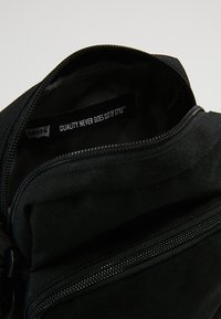 Pochette nera in tela con chiusura a zip. L'etichetta interna riporta la scritta "LA QUALITÀ NON PASSA MAI DI MODA™." Superficie testurizzata. Design minimalista.