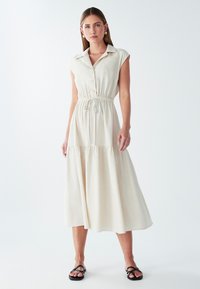 Hele beež kleit, ilma varrukateta, kaelusega, nuppude ees ja elastse vöökohtaga. Voogav kihiline seelik pehme tekstuuriga. Mustad sandaalid.