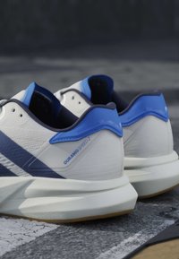 Chaussures de course blanches et bleues étiquetées « Duramo Speed 2 » posées sur une surface en béton bitumineux extérieure près d’une ligne blanche peinte.