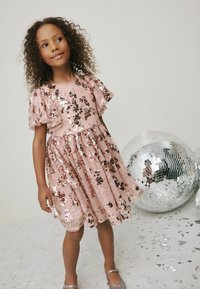 Rosa kjole med perler og blomsterdesign, korte flæsesleeves og en bølgende nederdel. Parret med glitrende sko og spejlvendte disco kugler.