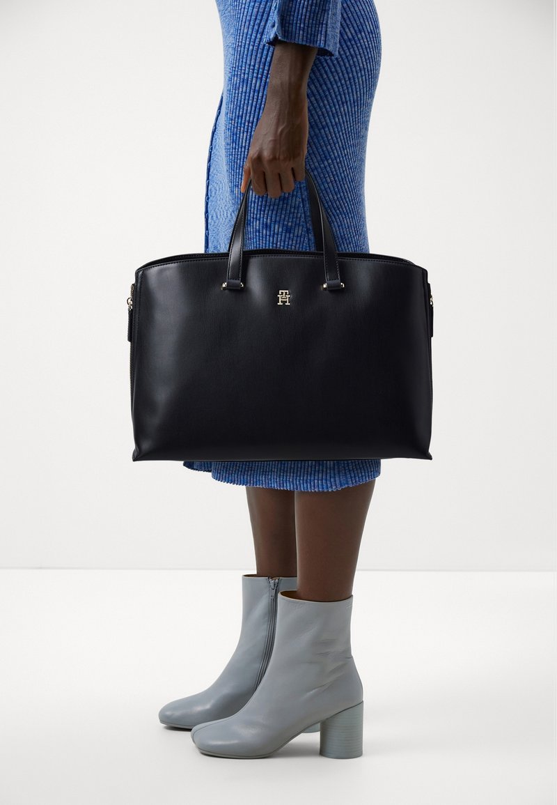 Personne en robe bleue tenant un grand sac à main en cuir noir, portant des bottines grises à talons blocs.