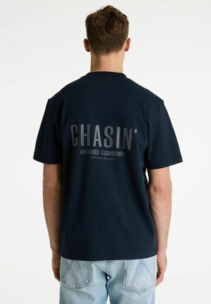 Man die wegkijkt, draagt een zwart T-shirt met 'CHASIN'' op de achterkant gedrukt en lichtblauwe spijkerbroek.