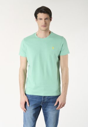 Uomo che indossa una t-shirt verde chiaro a maniche corte con un piccolo logo giallo sul petto e jeans blu, in piedi davanti a uno sfondo neutro.