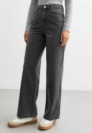 Jeans a vita alta e gamba larga in denim grigio scuro. Presentano tasche frontali e chiusura con bottone, abbinati a sneakers bianche e beige.