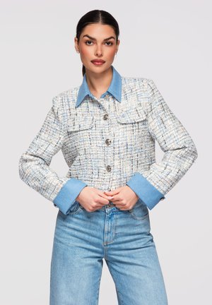 Žena v károvaném tvídovém saku s modrým denimovým límcem a manžetami, doplněném světle modrými džínami, stojící na světlém pozadí.