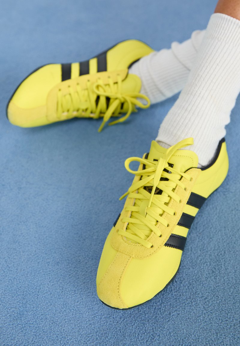 Sapatos deportivos amarelos brilhantes com riscas e atacadores pretos; apresentam um cabedal texturizado e detalhes em camurça. Usados com meias brancas caneladas.