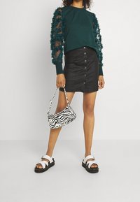 Vrouw draagt een donkergroene top met doorschijnende gerimpelde mouwen, een zwart minirokje met knopen, witte sandalen en houdt een handtas met zwart-witte zebra-print vast.