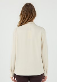 Beige Bluse mit langen Ärmeln und Kragen, glatte Textur und knöpfbaren Manschetten, mit einem leicht geschwungenen Saum hinten.