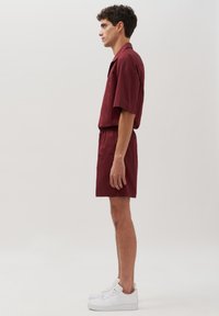 Camicia bordeaux a manica corta e pantaloni corti coordinati in un tessuto leggero, abbinati a sneakers bianche, indossati in una vista laterale.
