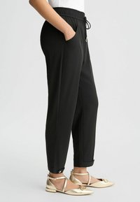 Schwarze tapered Hose mit elastischem Bund und Kordelzug, ausgestattet mit seitlichen Taschen und umgeschlagenen Säumen. Glattes Material mit leichtem Finish.