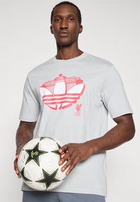 Γκρι κοντομάνικο μπλουζάκι με κόκκινο γραφικό ενός σταδίου και τρεις ρίγες στο στήθος, με λογότυπο LFC. Το μοντέλο κρατάει μία μπάλα ποδοσφαίρου.