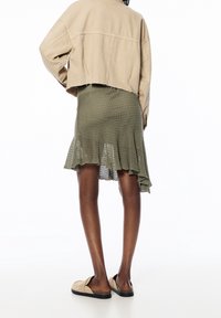 Beige cropped jack, olijfgroene geribbelde gebreide rok met een gerimpelde zoom, en lichtgekleurde instapschoenen met een gestructureerde zool.