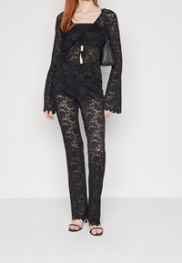 Svart spets tvådelad set bestående av en långärmad topp med knytdetalj och utsvängda ärmar, i kombination med åtsittande spetsbyxor.