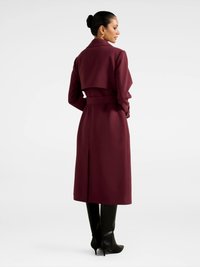 Bordoo pikk mantel jäigast materjalist, millel on lai krae, kinnitatud vöökoht ja seljalaputusdetail, koos põlvekõrguste mustade saapatega.