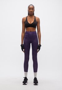 Femme athlétique debout, face à l'avant, portant un soutien-gorge de sport noir, un legging violet, des gants noirs, des chaussettes blanches et des baskets noires sur un fond blanc.
