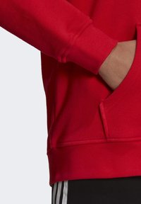 Röd sweatshirt med ribbad mudd och framficka. Materialet är mjukt med en slät yta. Synliga sömmar längs fållen.