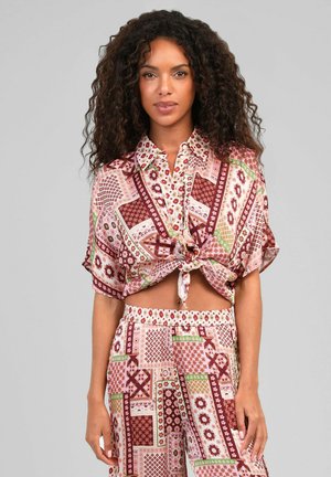 Femme aux cheveux bouclés portant un haut à motifs assortis noué à la taille et un pantalon taille haute à imprimé géométrique rose, rouge et vert.
