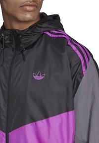 Chaqueta de nylon negra y morada con cierre frontal, capucha, tres franjas moradas a lo largo de los hombros y un logo sutil en el pecho.