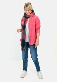 Roze button-up shirt over een wit T-shirt, blauwe jeans en een veelkleurige franjesjaal. Heldere sneakers maken de outfit compleet.