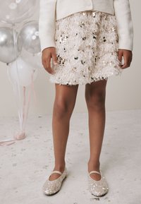 Pufasta, svijetloružičasta suknja s šljokicama, uparena sa sjajnim bež baletankama s remenčićem. U pozadini su baloni i raspršeni konfeti.