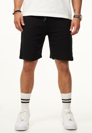 Schwarze Shorts aus strukturiertem Material, mit elastischem Bund und Kordelzug. Kombiniert mit weißen Turnschuhen und weißen Socken mit schwarzen Streifen.