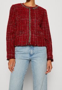 Vrouw in een rode, gemêleerde tweedjasje met kettingdetails, over lichtblauwe high-waisted jeans, staand voor een effen achtergrond.