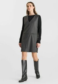 Grijze krijtstreep mouwloze jurk met een V-hals, zijzakken, gedragen over een zwart longsleeve; gecombineerd met zwart leren kniehoog boots.
