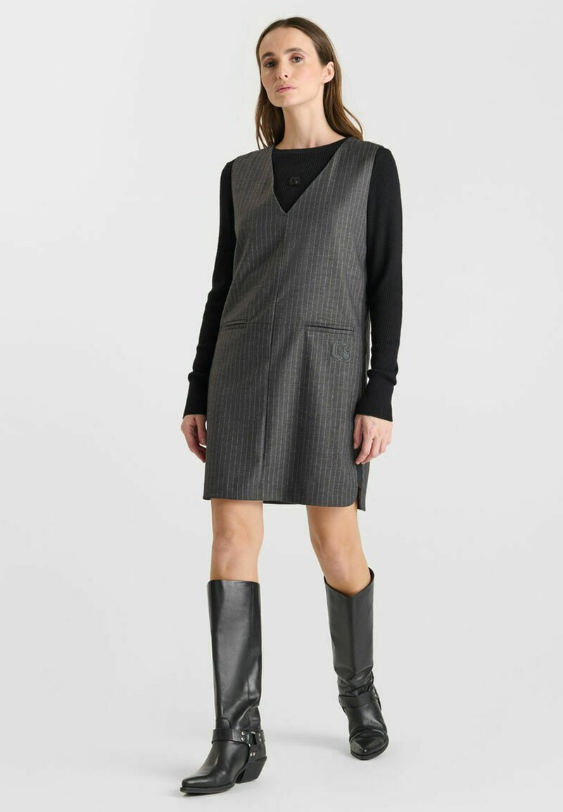 Grijze krijtstreep mouwloze jurk met een V-hals, zijzakken, gedragen over een zwart longsleeve; gecombineerd met zwart leren kniehoog boots.