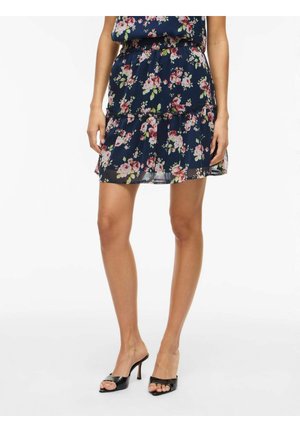 Femme portant une mini-jupe à volants fleurie bleu marine associée à des chaussures à talons hauts noires ouvertes à l’avant, debout sur un fond blanc.