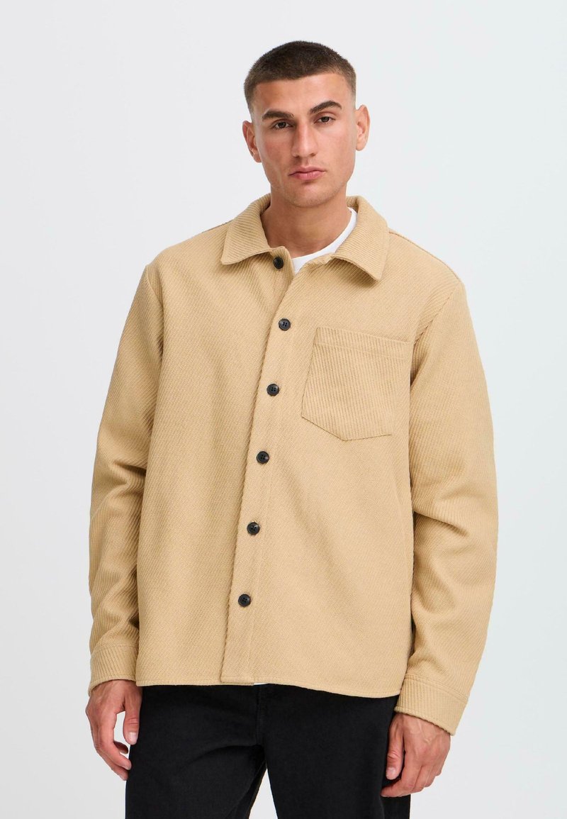 Beige corduroy shirt jacket with a buttoned front, single chest pocket, and black buttons. Features a textured fabric and pointed collar.