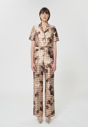 Femme portant une tenue deux-pièces beige à carreaux vichy avec des imprimés floraux, un haut à manches courtes boutonné, un pantalon à jambes larges et des chaussures métalliques à bouts pointus.