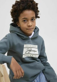 Sudadera gris con un parche de logo con el texto "JackJones", puños acanalados y un bolsillo tipo canguro, llevada sobre una camisa blanca.