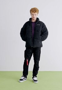 Zwarte fleece pufferjack met hoge kraag, gecombineerd met zwarte cargobroek met een rode ritssluiting en zwart-witte sneakers.