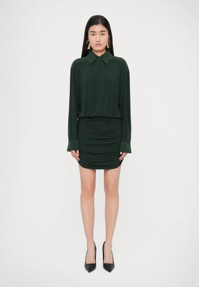 Dark green blouse with a collar and long sleeves, paired with a ruched mini skirt in the same color. Model wearing black pointed heels.